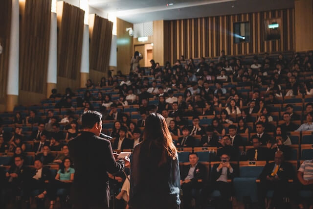 Two people with their backs to the camera speak to an audience in an auditorium.