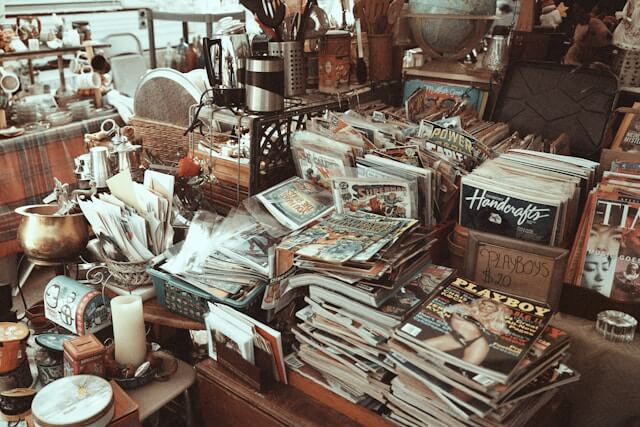 A messy table is covered with magazines, random art and kitchen supplies, and trinkets.