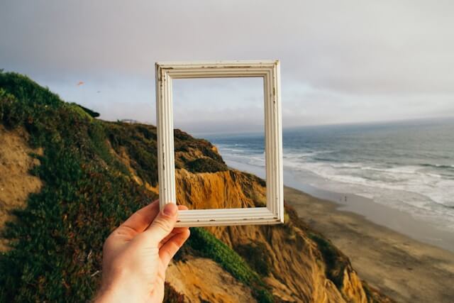 A hand holds a picture frame in front of the camera, in the background is a Californian coastline.