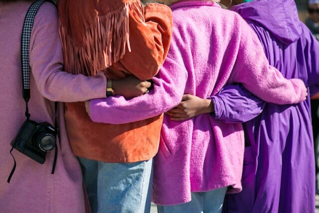 Four women wearing bright colours stand with their arms around each other and their backs to the camera.