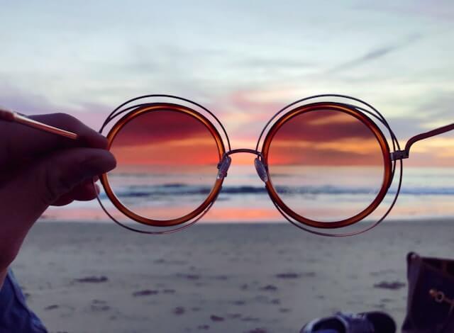 Sunglasses are held in front of the camera and a sunset on the beach looks more saturated inside them.