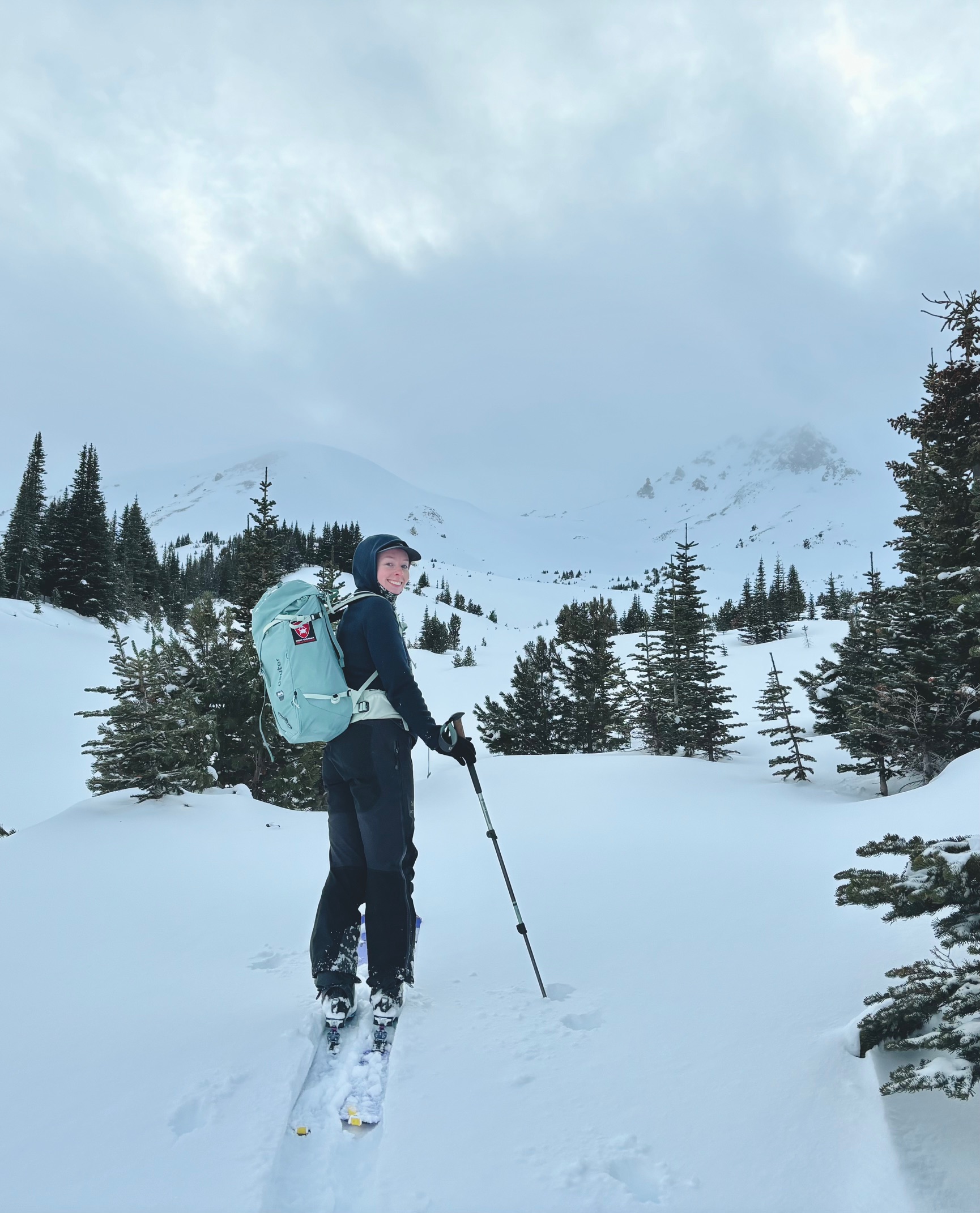 A photo of a woman looking back at the camera as she walks uphill on backcountry skis.