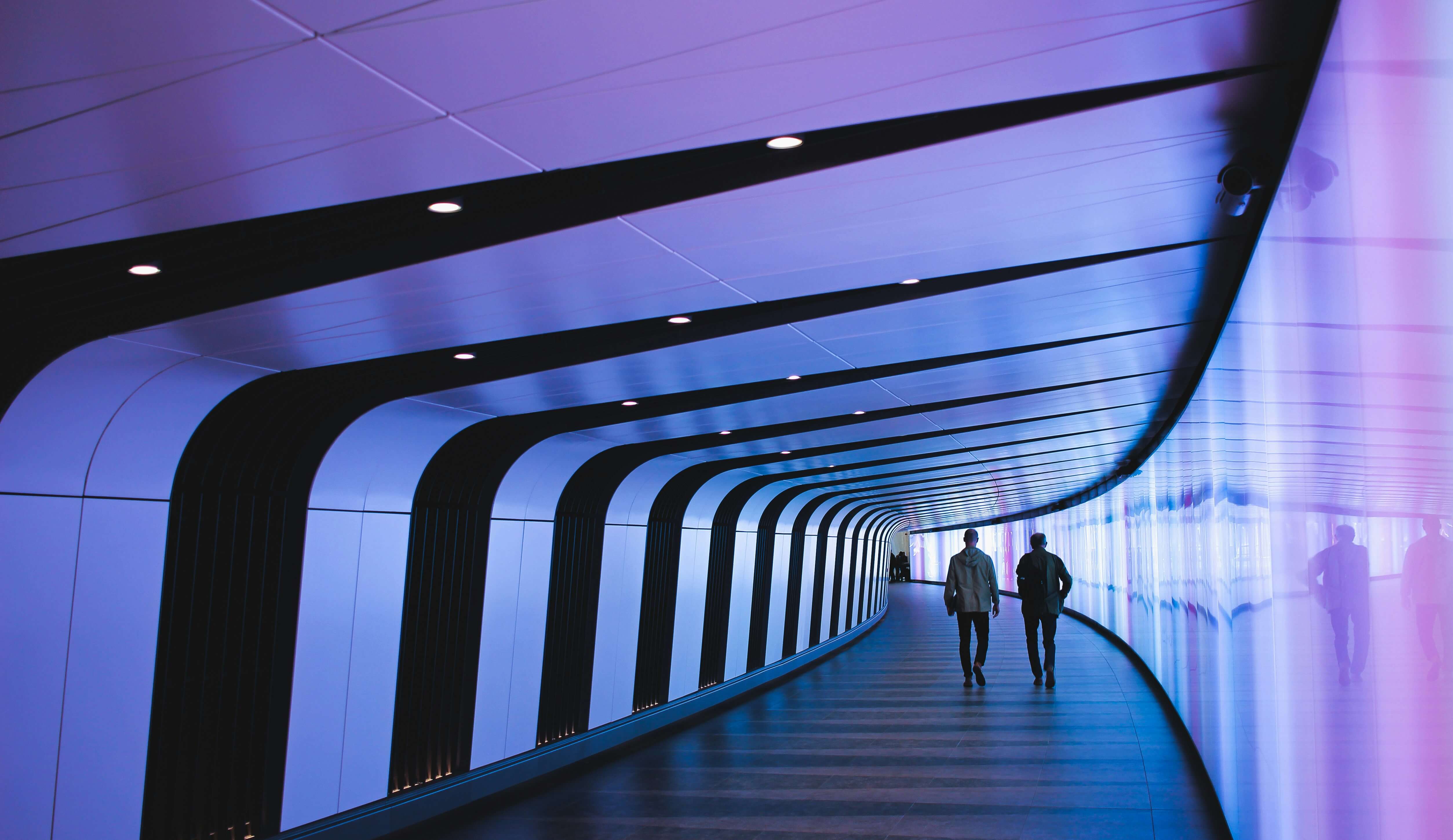 two men walk down a curving hallway filled with purple and white lighting.