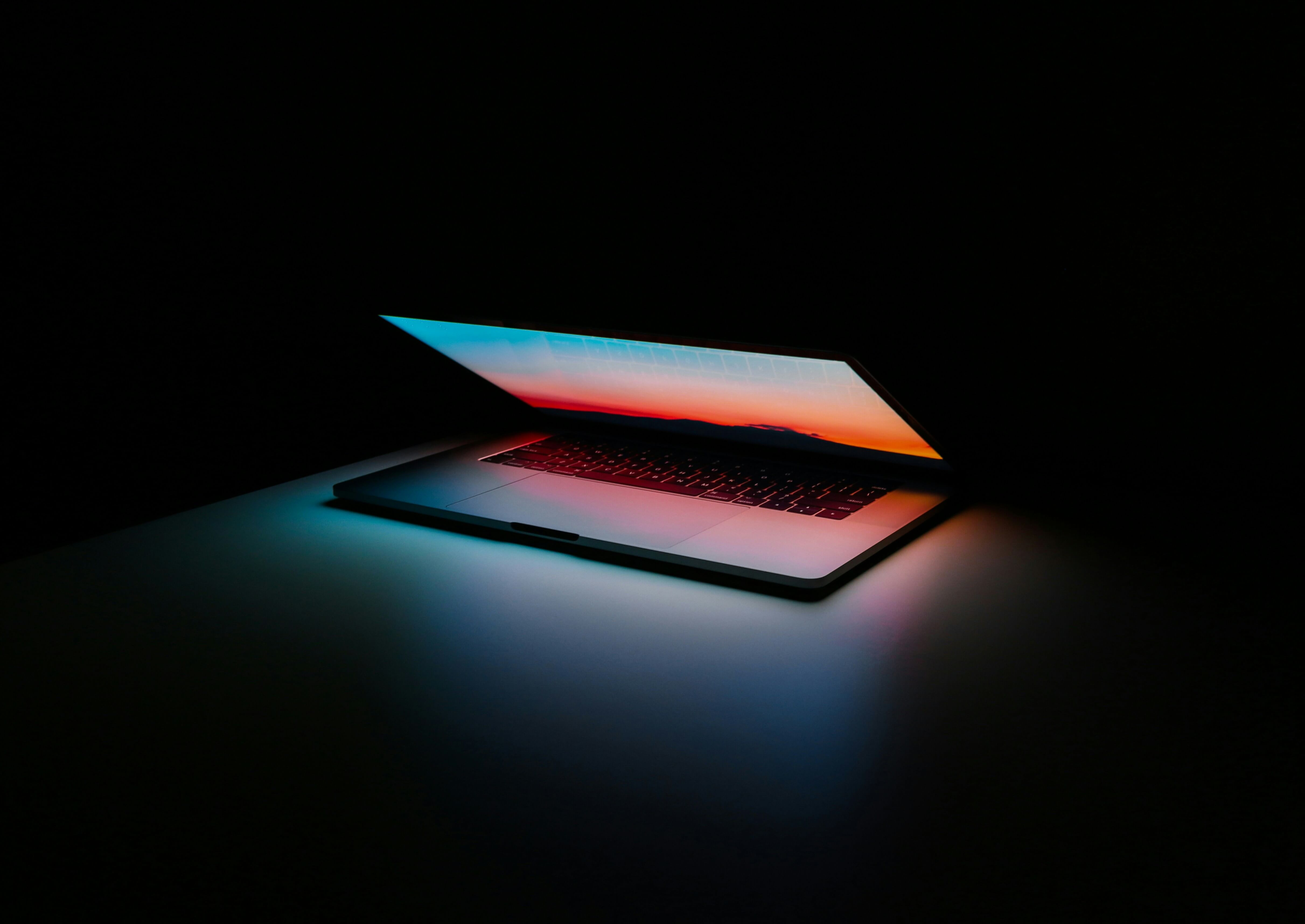 A laptop sits halfway open in the dark, casting glowing lights from its screen around it.