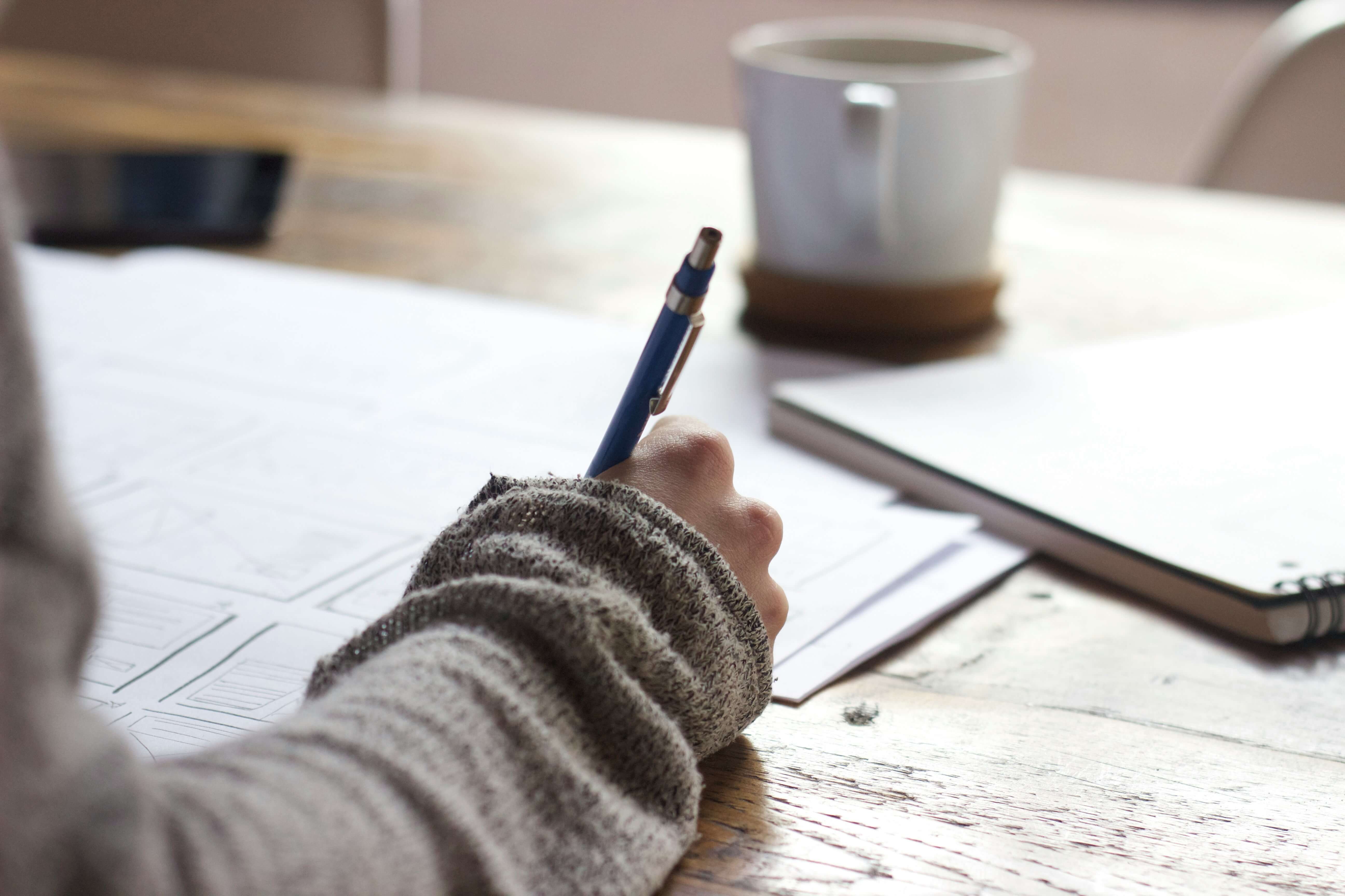 A woman's arm writes with a pen on a piece of paper. A coffee mug sits in front of her on the table.